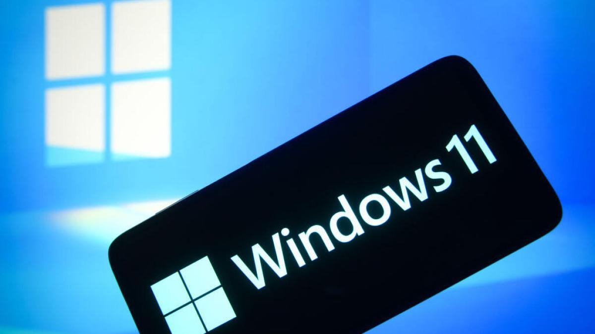 A phone with a black screen and light blue Windows 11 logo is in the foreground. A light blue room with a white Windows logo is in the background.