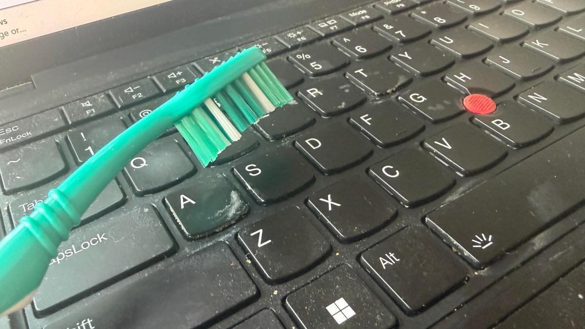 A disgusting grimy ThinkPad keyboard is in the background while someone holds a clean, new teal toothbrush in front of it.