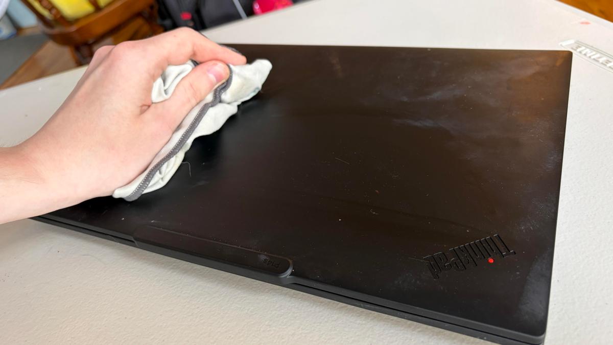 A black ThinkPad computer is covered in dust and grime while a hand holding a microfiber cloth wipes it down.