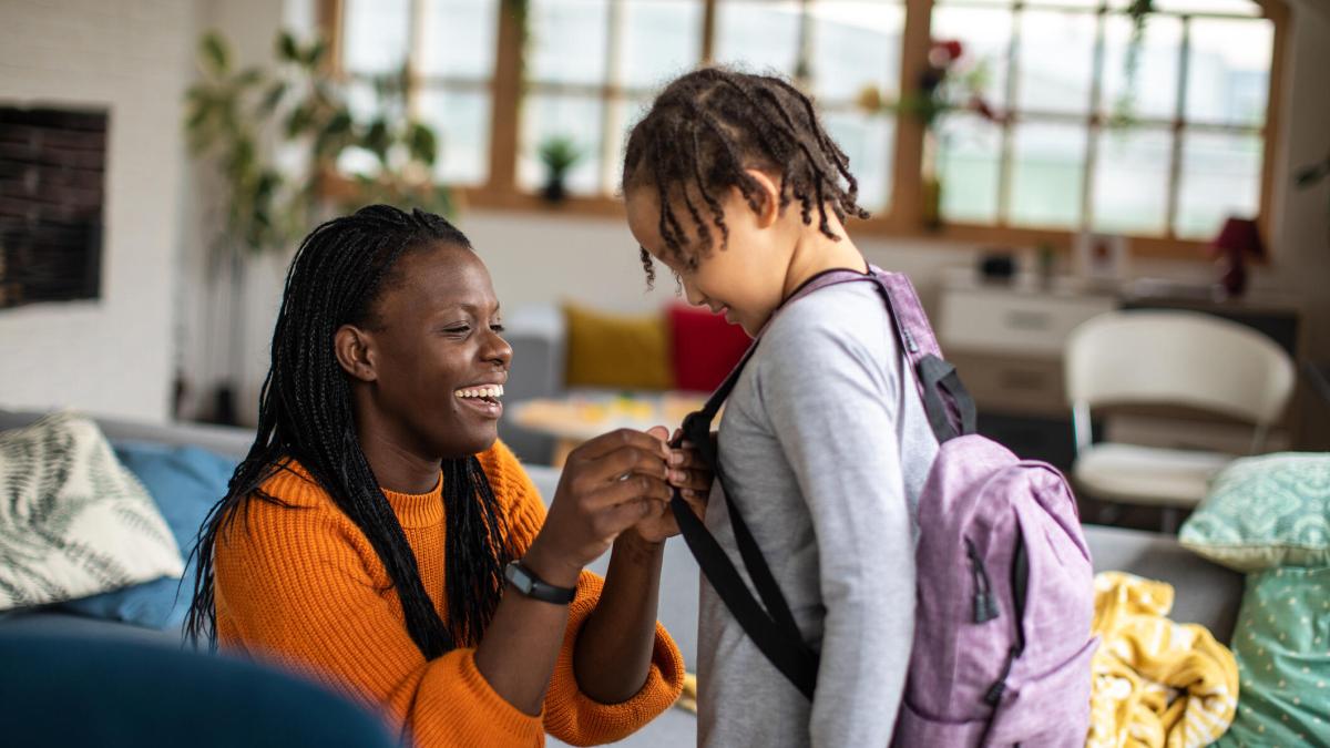 A smiling mother adjusts backpack straps for a young child in a living room.