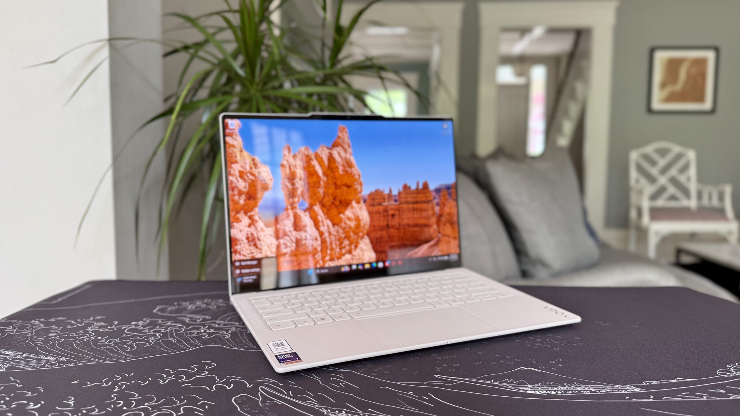 Yoga Slim 7i Ultra Aura Edition Gen 11 sits on a table with a black and white mat underneath it. A living room with some greenery is in the background.